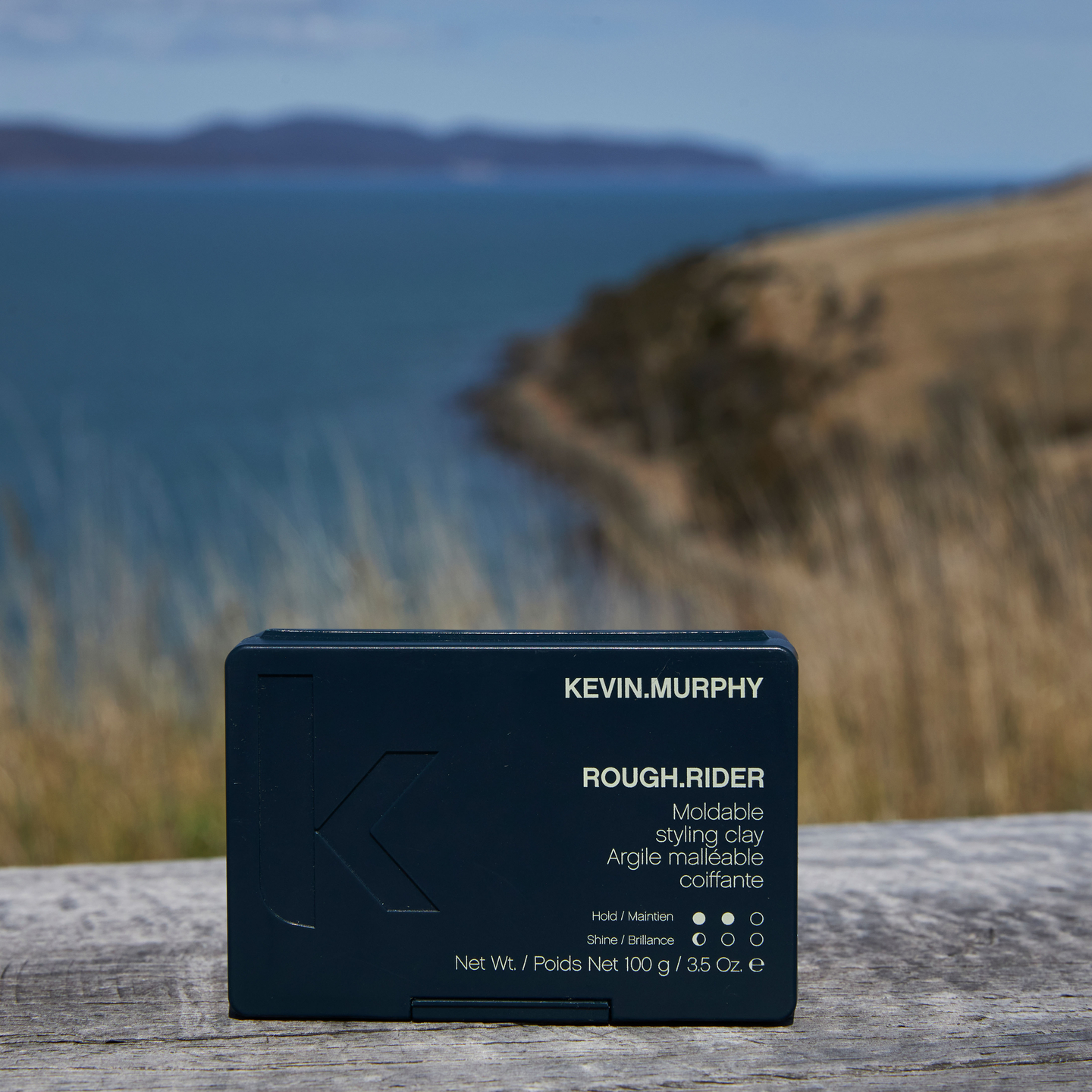 Outdoor image of KEVIN.MURPHY ROUGH.RIDER styling clay packaging, displaying the product's name and attributes like hold and shine level on the label, set against a blurred natural backdrop.